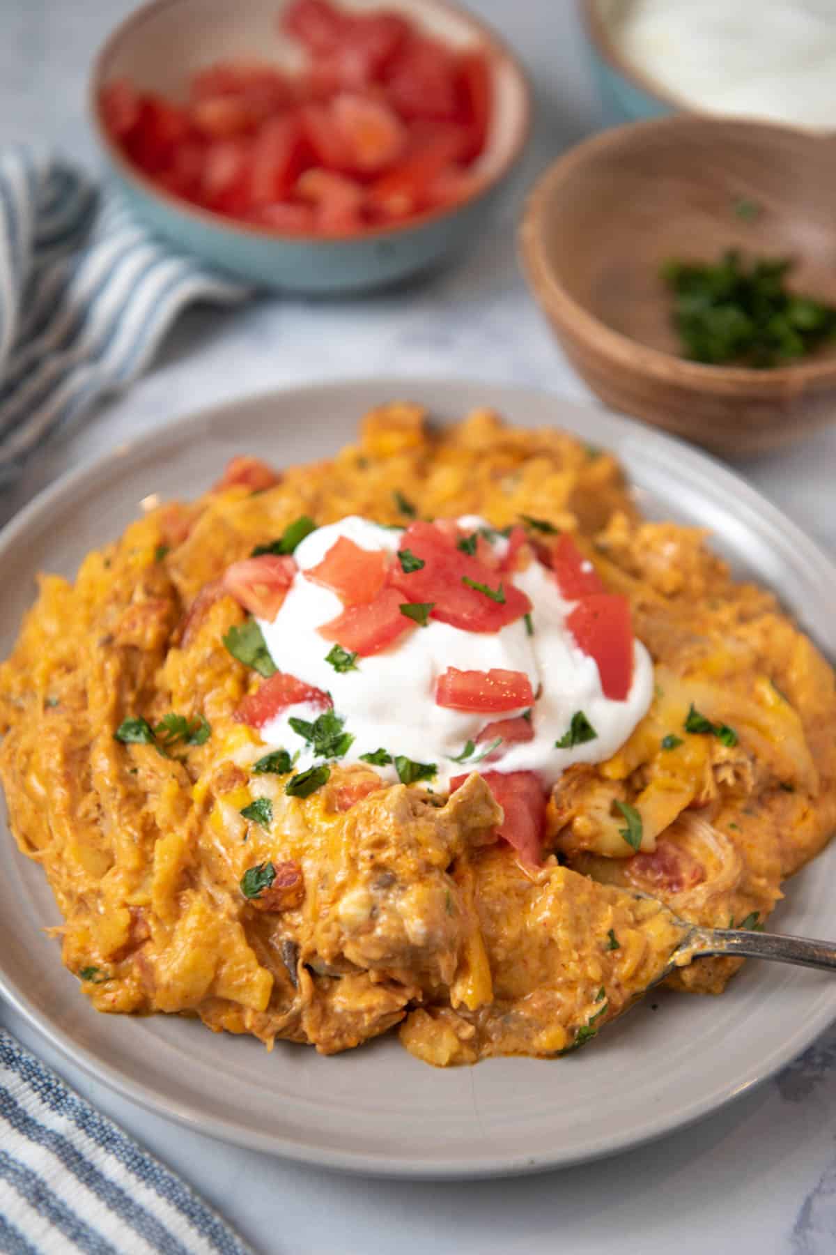 serving of Crock Pot chicken enchilada casserole topped with sour cream and tomatoes on gray plate, with forked bite