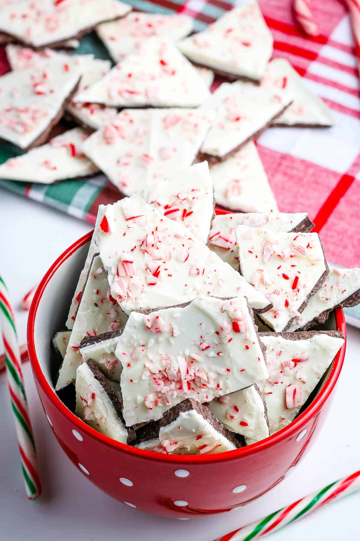 pile of white chocolate peppermint bark in red bowl and on plaid Christmas towel on white countertop with candy canes