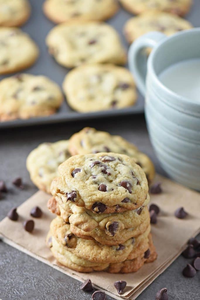 chocolate chip cookies stacked on brown paper bag with chocolate chips and blue mug of milk