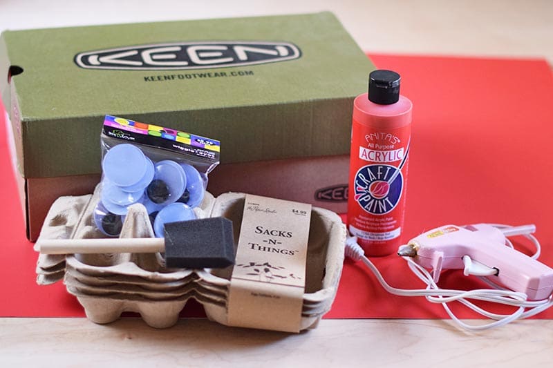 craft supplies for making a dinosaur Valentine box, including a shoebox, hot glue gun, paint, poster board, an egg carton, wiggle eyes, and a sponge paint brush