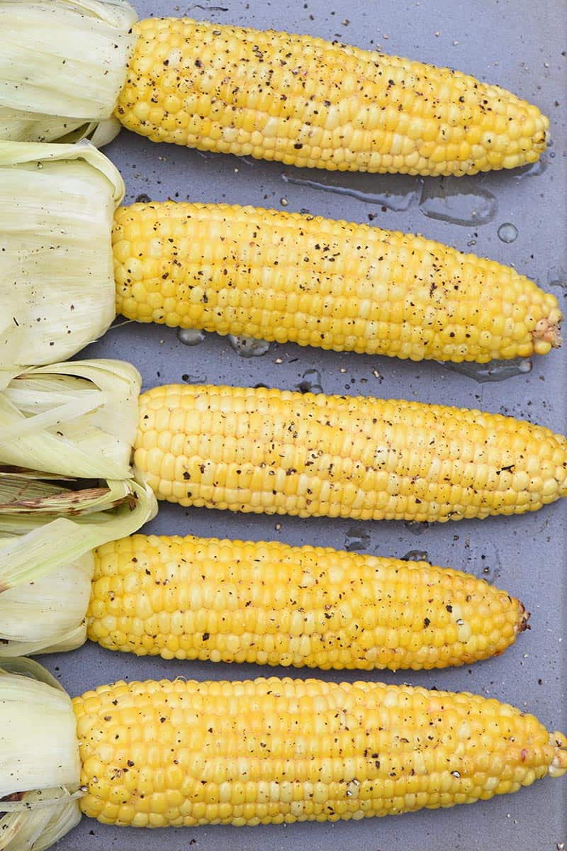 grilled corn in the husk on a cookie sheet with husks removed, seasoned with honey butter, salt, and pepper