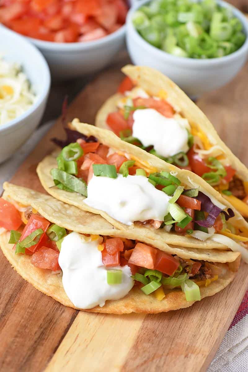best ground beef tacos ever on a wooden cutting board with colorful toppings like cheese, tomatoes, and green onions