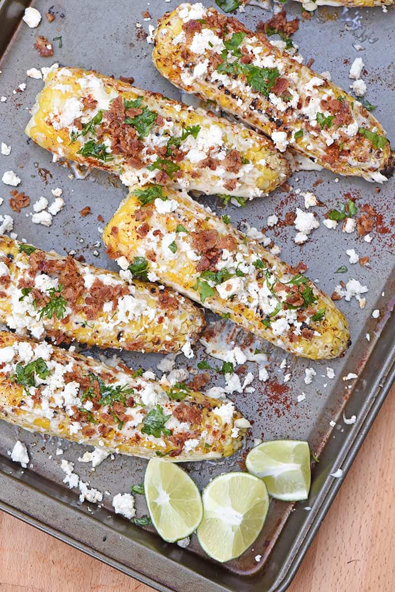 grilled Mexican corn on baking sheet with lime wedges and bacon crumbles