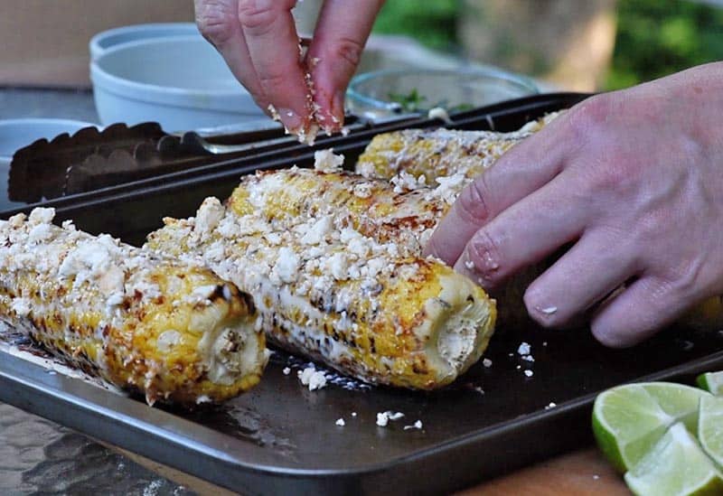 crumbling Feta cheese onto grilled corn on the cob for Mexican corn recipe