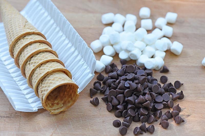 ingredients for campfire s'mores recipe, including sugar cones, Ghirardelli chocolate chips, and mini marshmallows on a cutting board