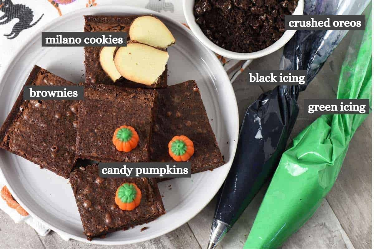 Graveyard brownies ingredients on wooden countertop, including brownie mix brownies, Milano cookies, green and black icing, Oreo Cookies, and candy pumpkins