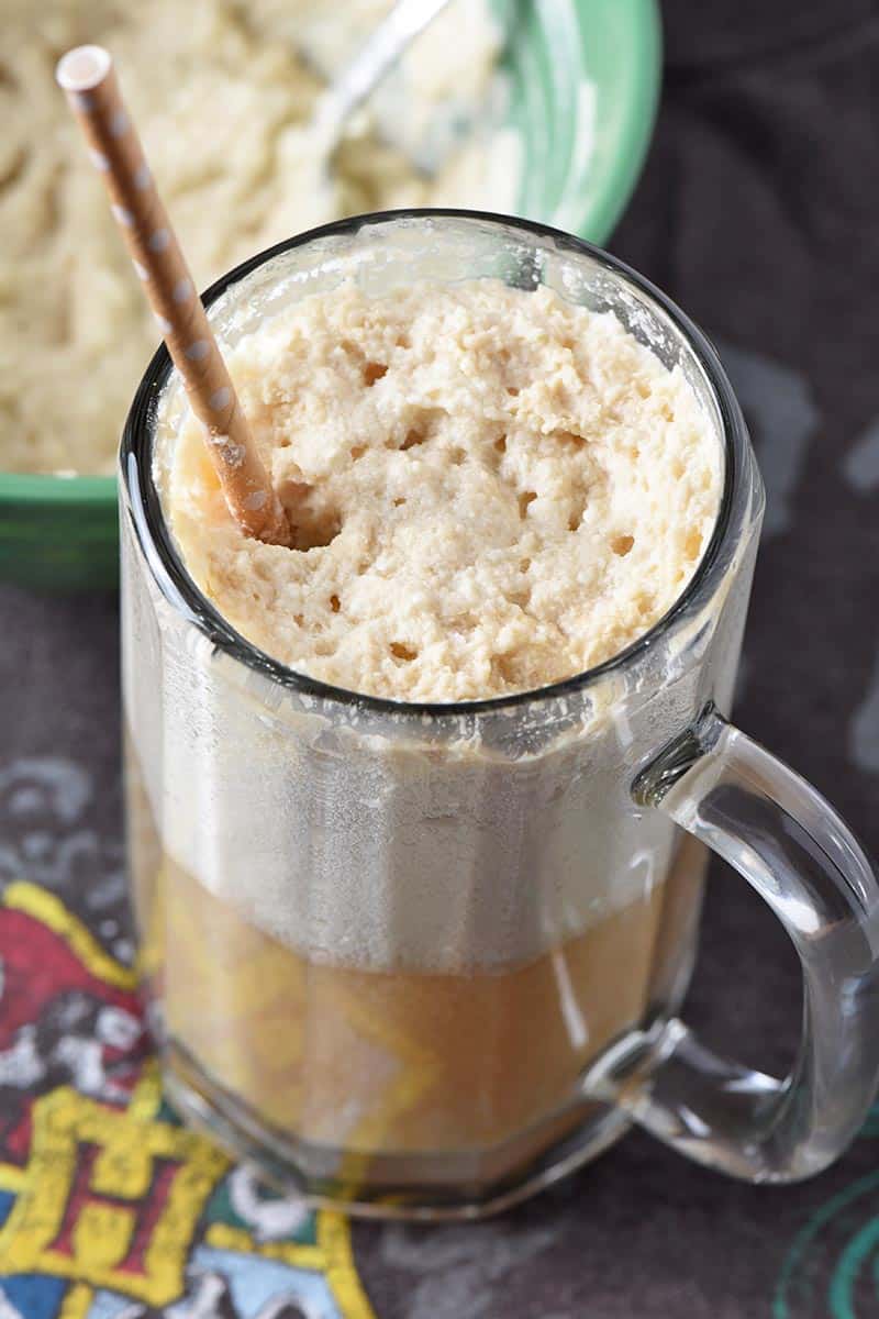 Tall glass mug of homemade butterbeer with frothy whipped cream topping and a paper straw on a Hogwarts shirt