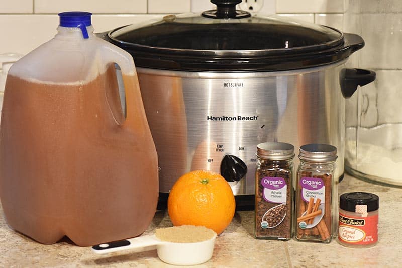 ingredients for making mulled cider, including apple cider, oranges, brown sugar, cinnamon sticks, allspice, and whole cloves