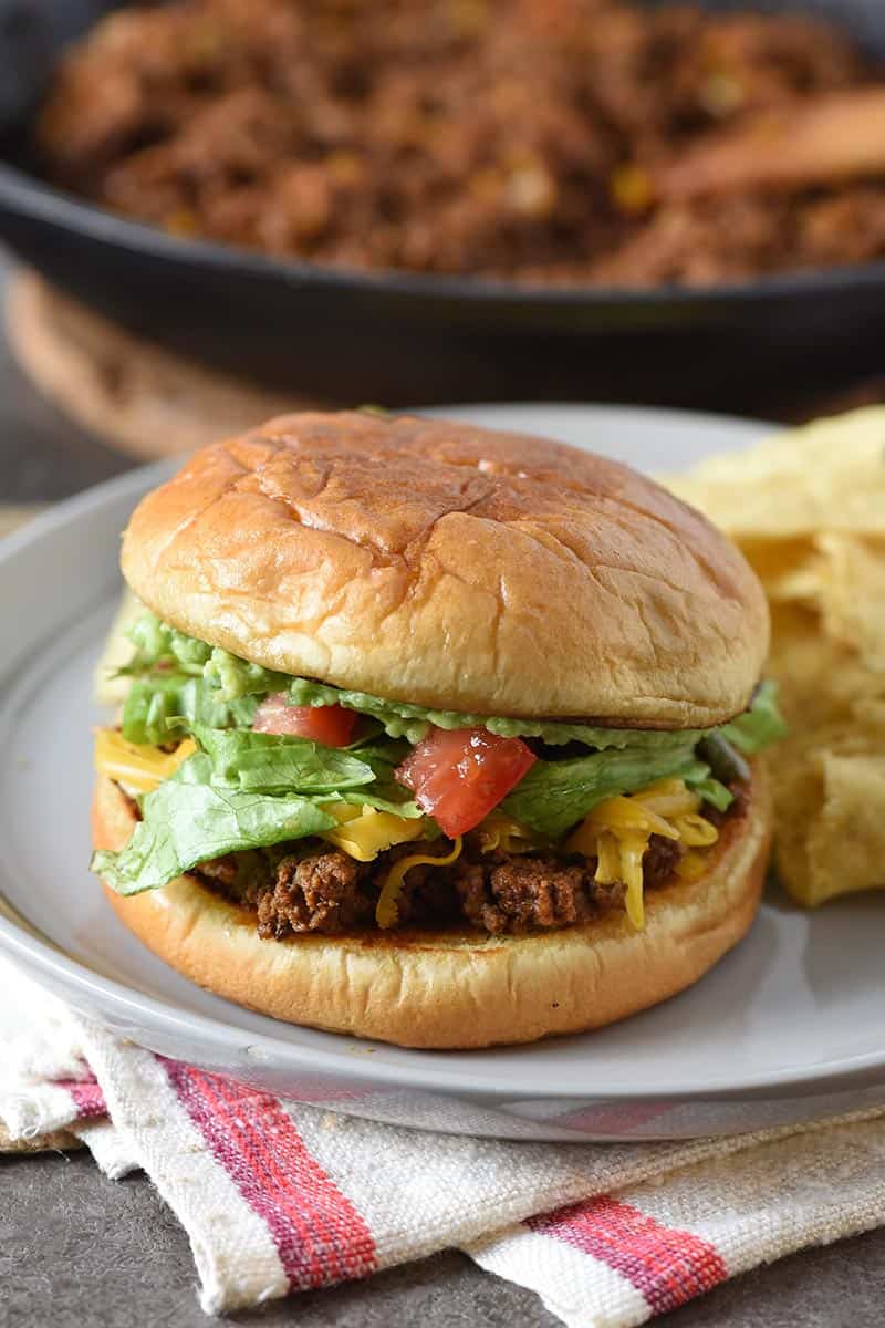 taco sloppy joes on toasted buns served on gray plate with tortilla chips and cast iron skillet of juicy burger meat behind it