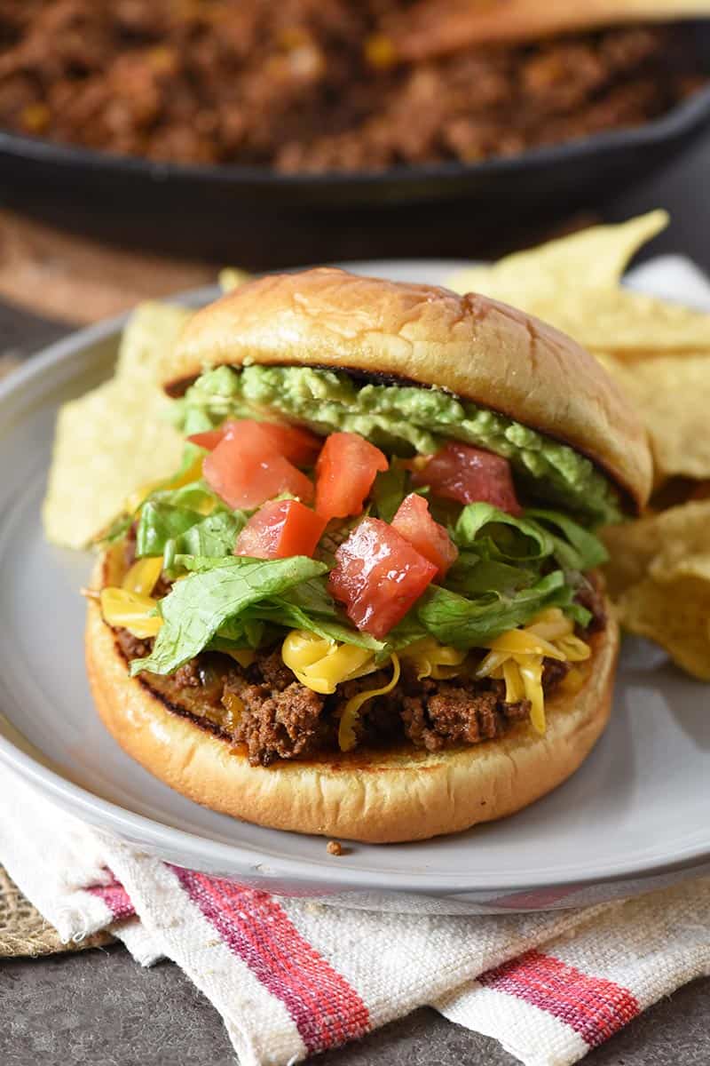 taco ingredients like avocado, tomatoes, lettuce, and shredded cheddar piled on sloppy joes on gray plate with tortilla chips