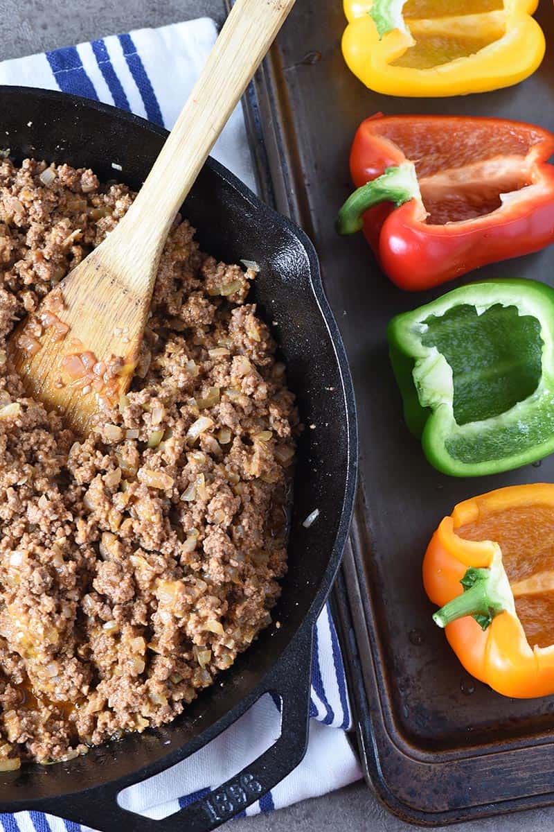 ingredients for sloppy joe stuffed peppers, including ground beef in cast iron skillet with wooden spatula and bell peppers on baking sheet