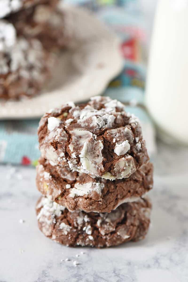 stack of chocolate crinkle cookies on white countertop with plate of Christmas cookies and milk