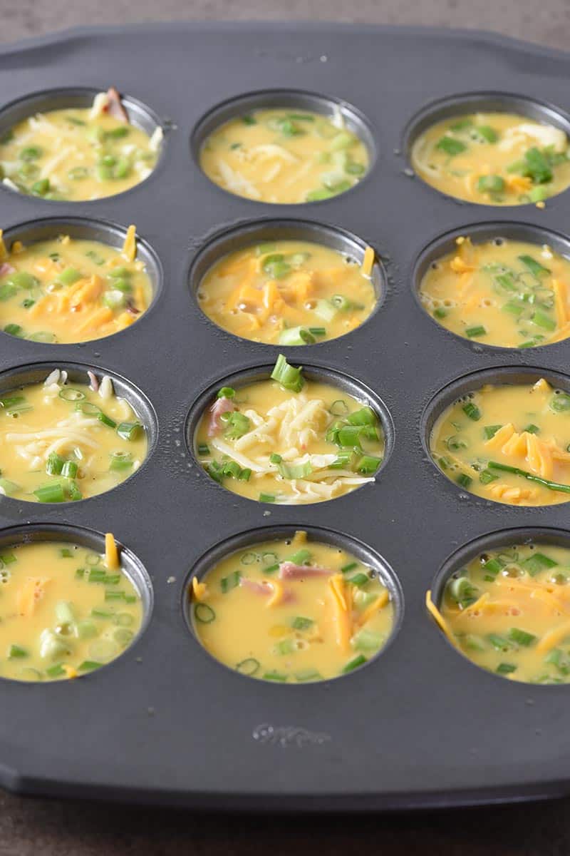 raw eggs, ham and cheese, and green onions in muffin pan ready to bake into egg muffins