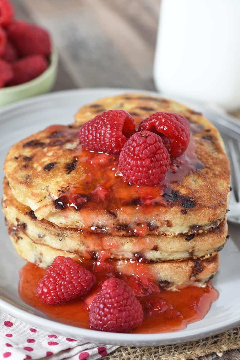 heart shaped pancakes with chocolate chips stacked on gray plate, slathered with raspberries and strawberry jam