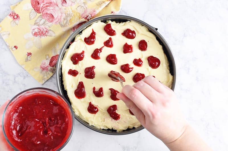 spooning dollops of raspberry sauce for cheesecake onto batter in baking pan