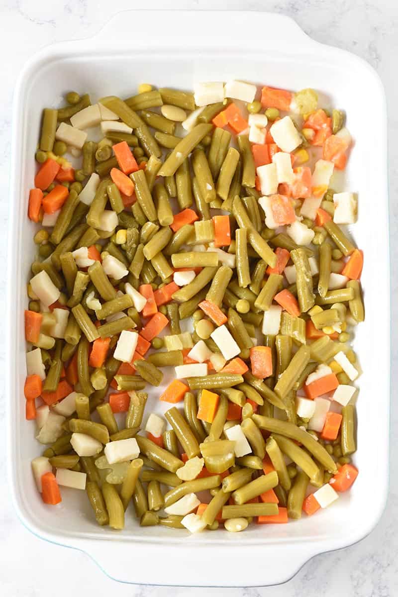 layering canned green beans and mixed vegetables to make tater tot casserole with veggies in blue and white casserole dish