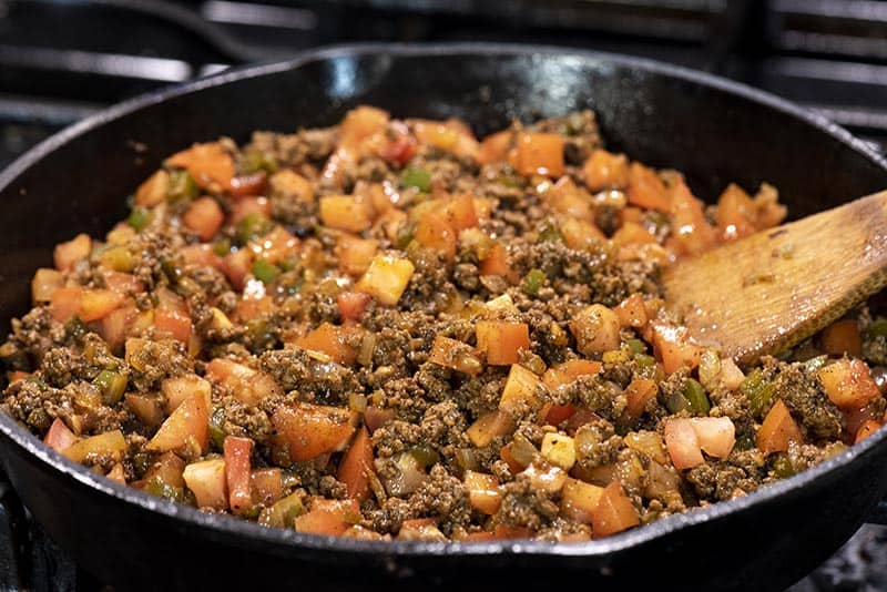 cooking ground beef mixture for taco tater tot hotdish in cast iron skillet with wooden spatula