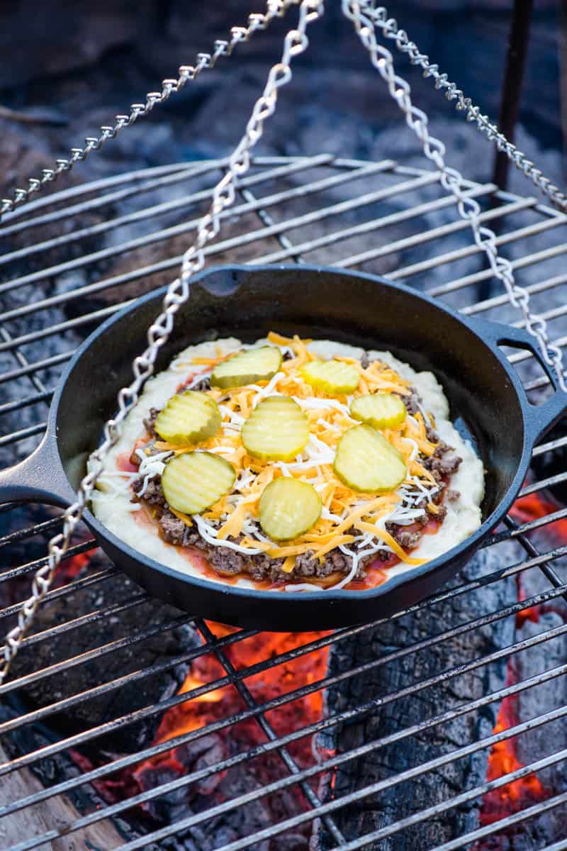 cooking hamburger pizza on a campfire in a cast iron skillet, using a tripod grill