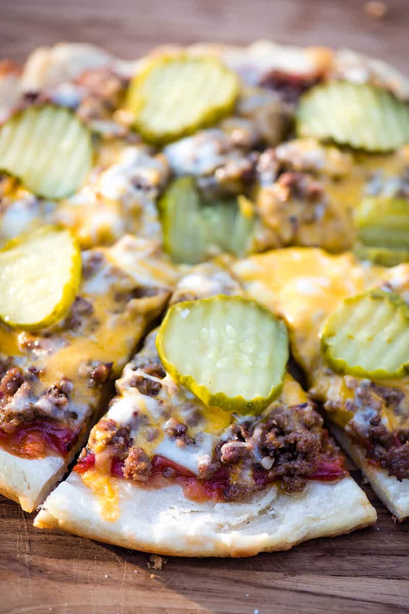 sliced hamburger pizza with pickles and melted cheese on a wooden cutting board