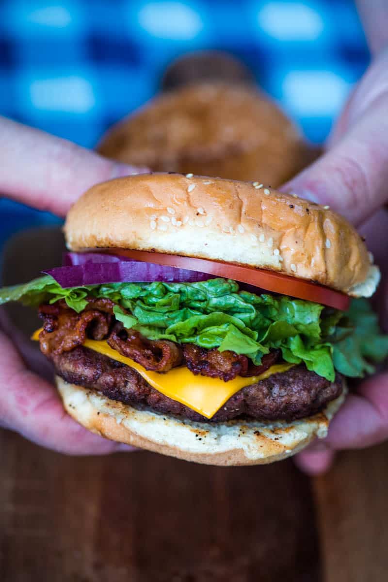 hands holding bacon burger over wooden cutting board on picnic table