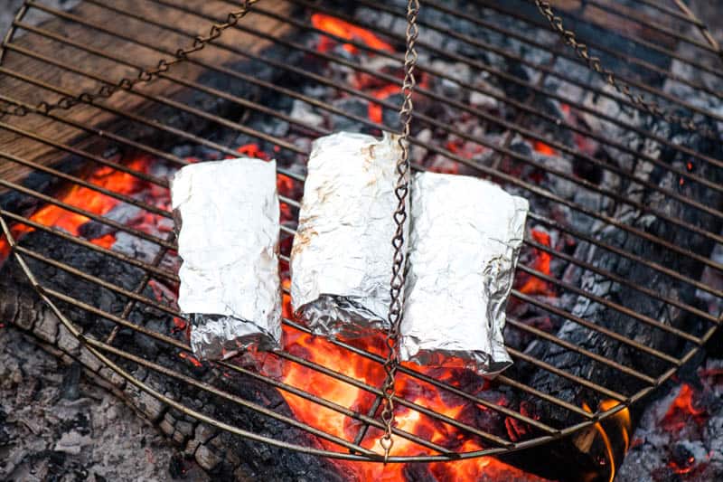 grilling camping breakfast burritos in heavy duty foil on a tripod grill over the campfire
