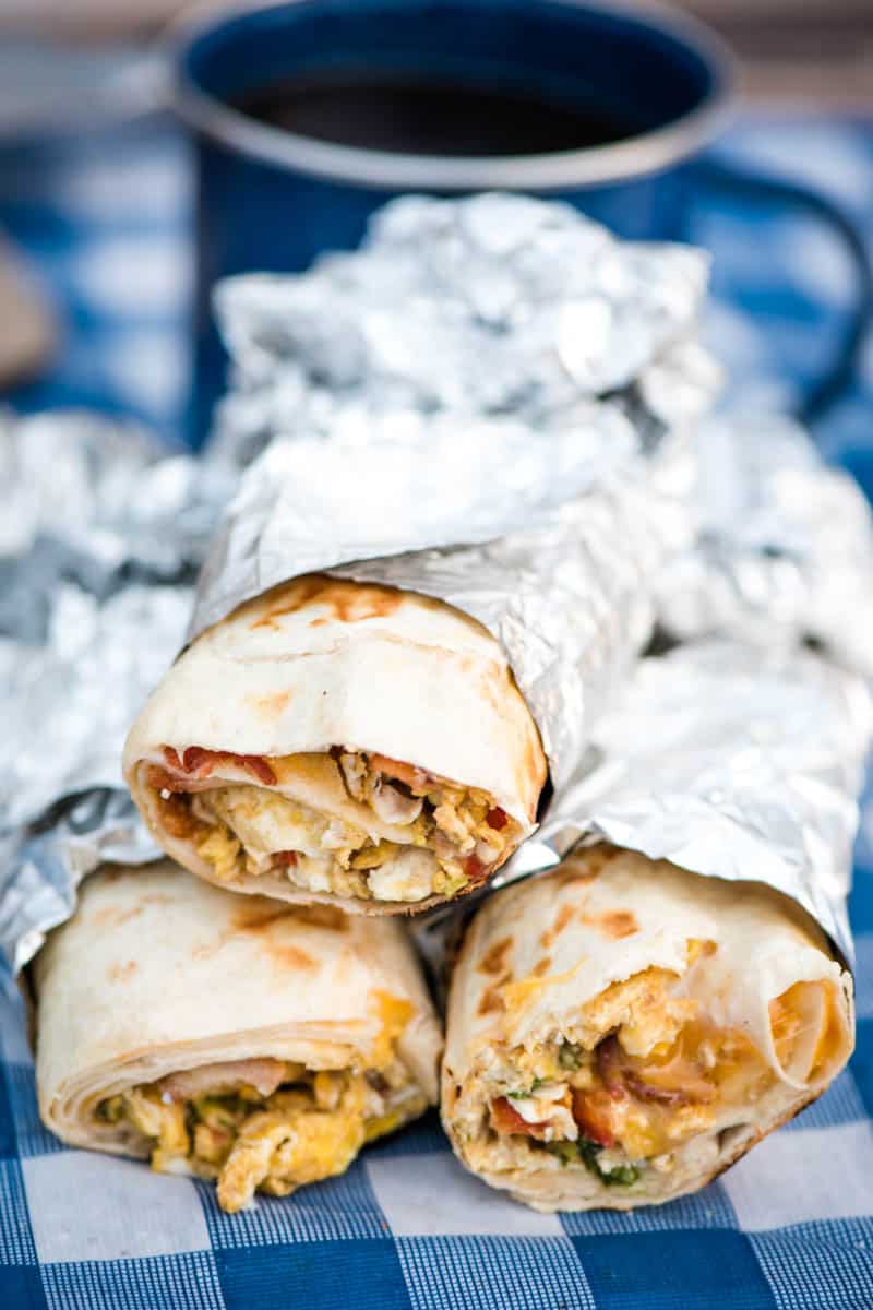 stack of make-ahead camping breakfast burritos, wrapped in foil, on blue gingham picnic table
