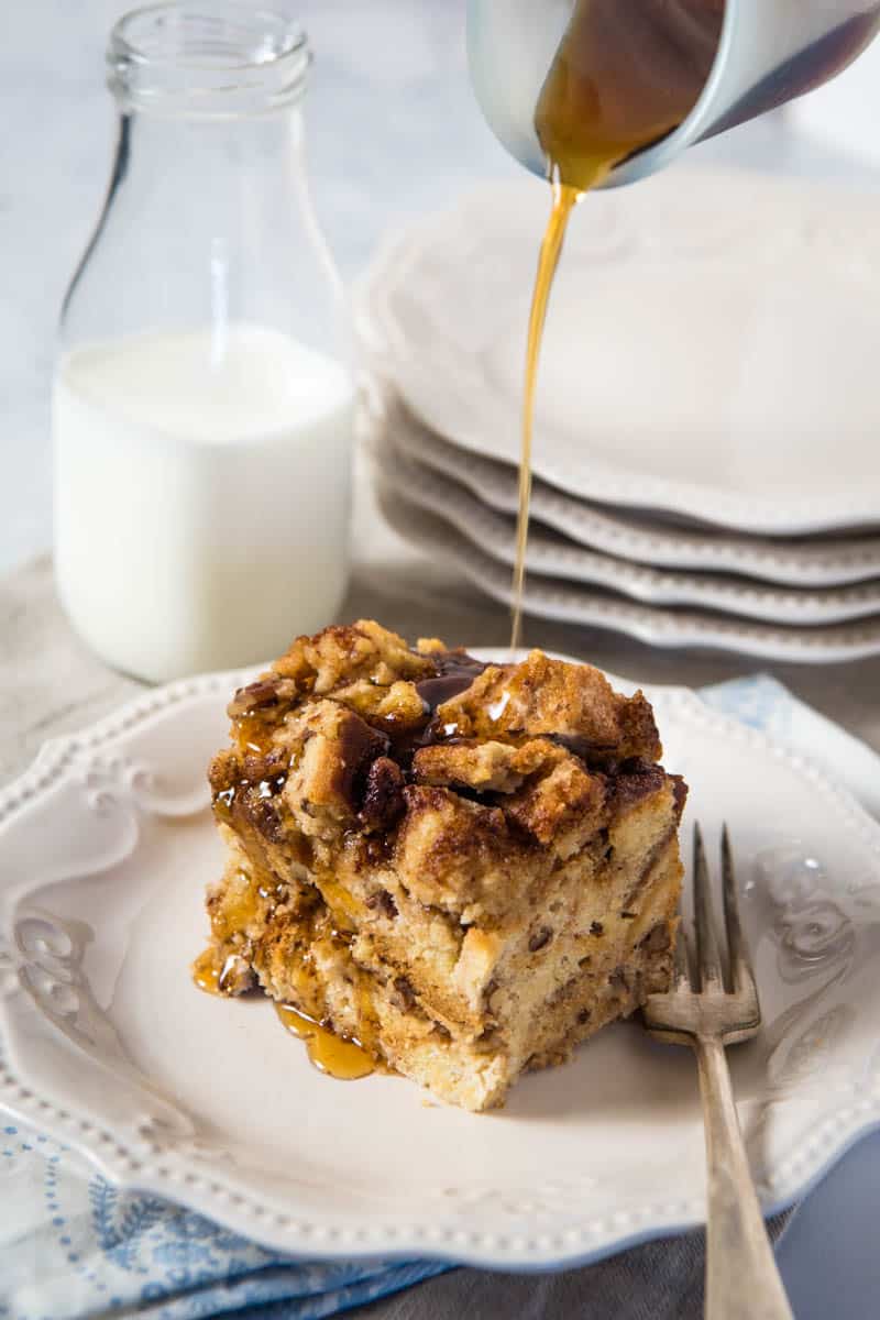 pouring maple syrup over slice of gluten-free French toast casserole on white plate with fork, glass of milk in background