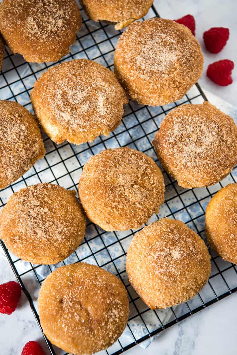 Bisquick cinnamon sugar muffins sitting on wire rack over blue/white flowered linen on white countertop, with fresh red raspberries