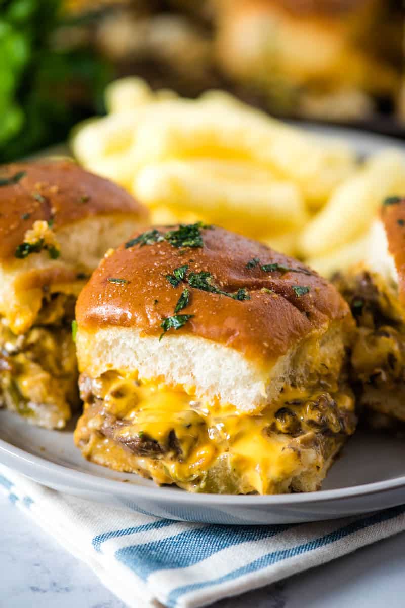Cheese steak sliders with Cheez Whiz, sprinkled with Italian parsley on gray plate, on blue and white striped tea towel on white marble countertop