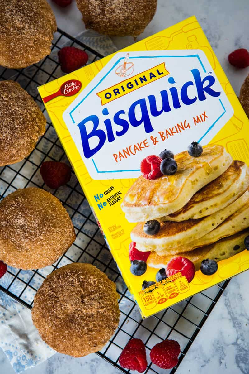 cinnamon Bisquick muffins with Bisquick mix on black wire rack over white countertop with blue and white flowered linen, with fresh red raspberries on rack and countertop