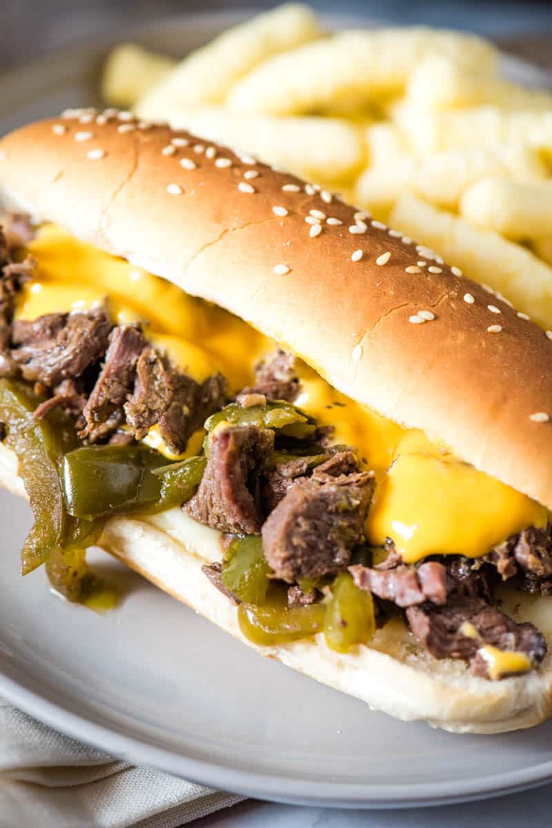 close up CrockPot Philly cheesesteak sandwich on hoagie bun with sirloin steak, onion, green bell peppers, and provolone cheese topped with Cheez Whiz, on gray plate with white cheese puffs