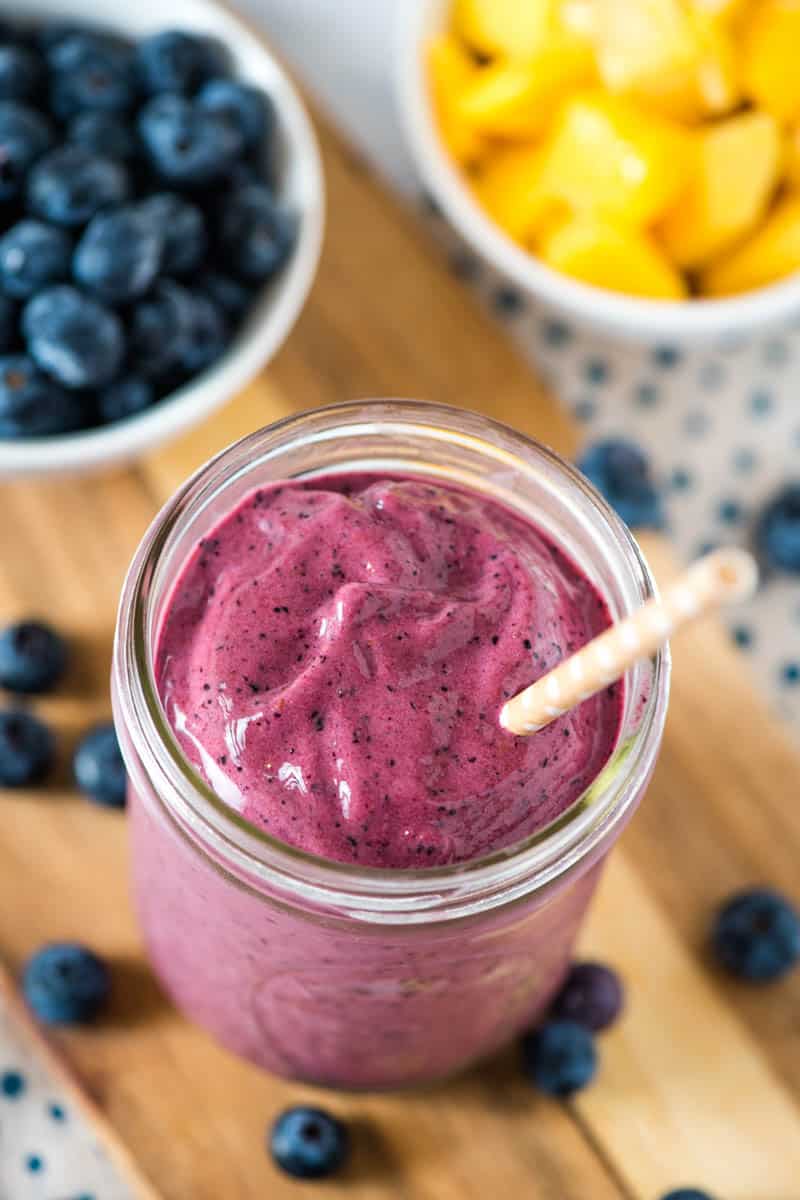 mango blueberry banana smoothie in mason jar with brown paper straw, on wood cutting board with blueberries and mango in small white bowls