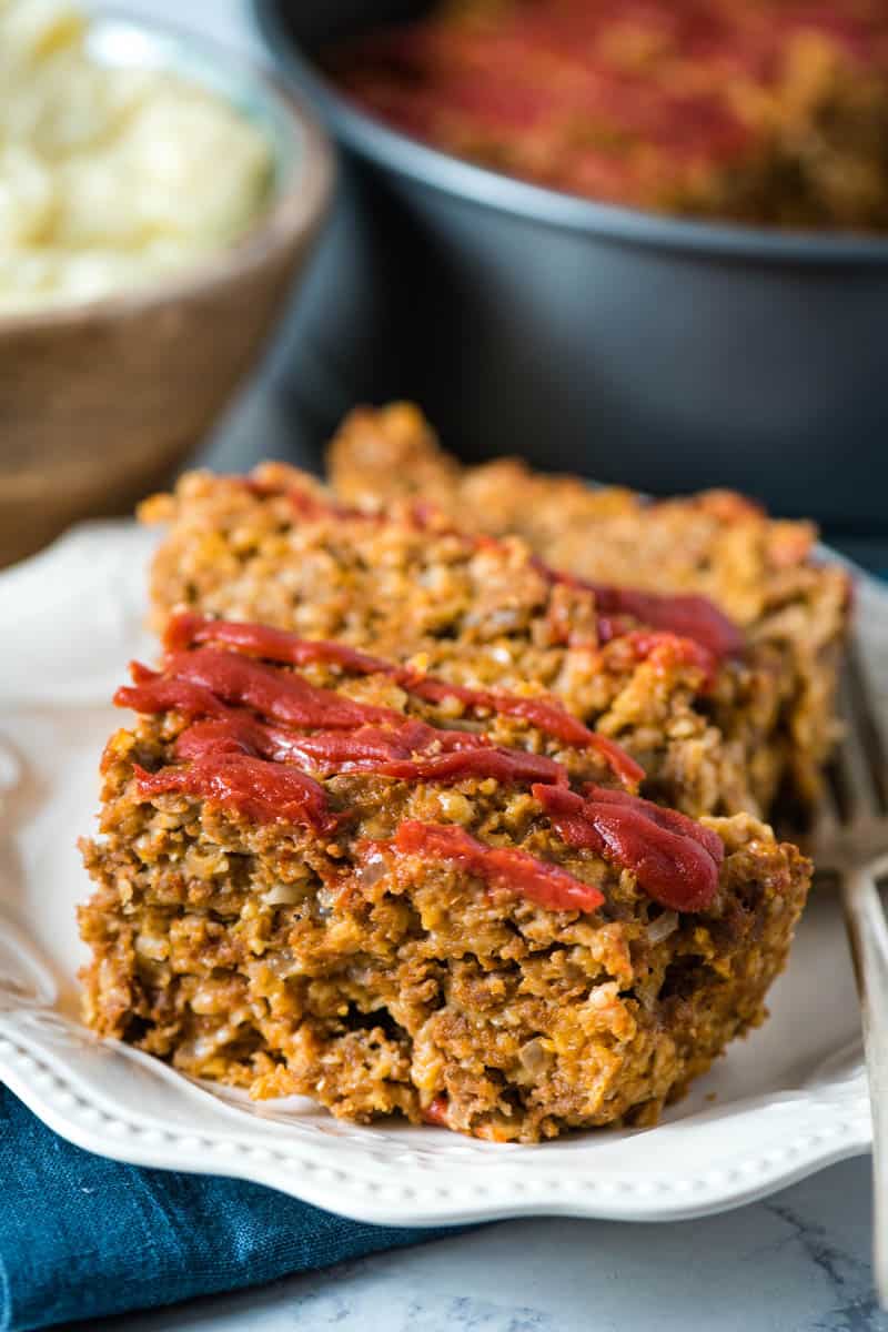 ketchup-topped meatloaf on white plate with fork and mashed potatoes in wooden bowl behind plate