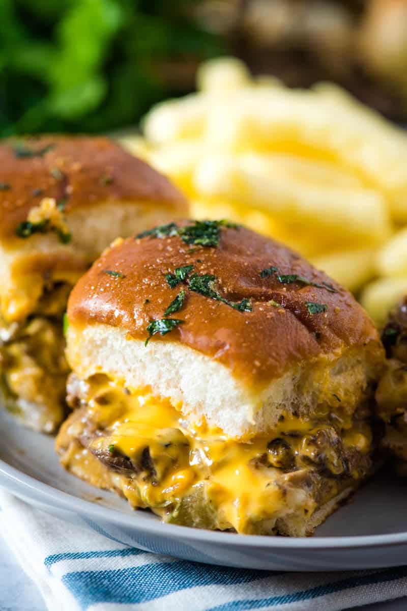 Philly cheese steaks on Hawaiian Rolls  brushed with garlic butter and Italian parsley, topped with Cheez Whiz, on gray plate with blue and white striped towel