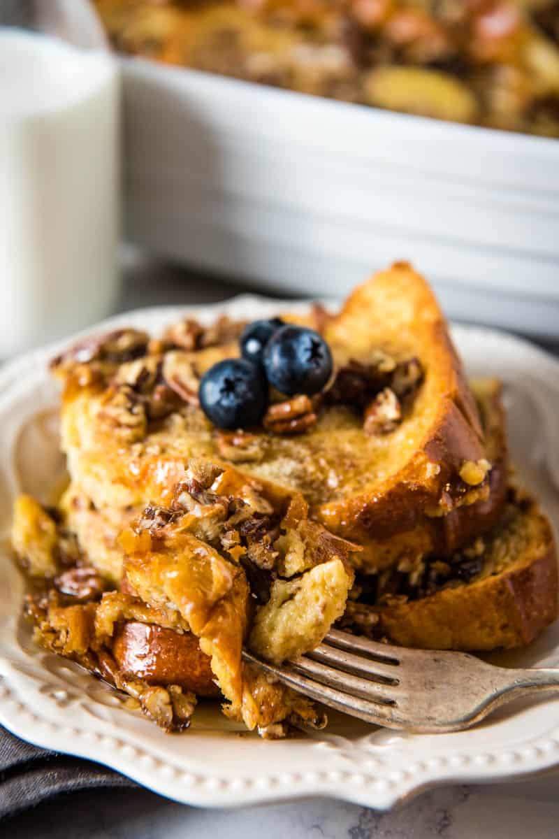 bite of brioche French toast casserole on fork with French toast, smothered in maple syrup, behind on white plate