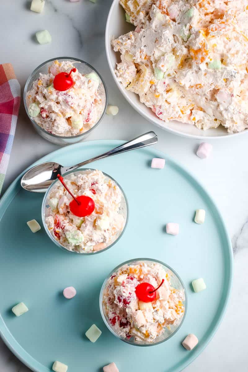 above large bowl of ambrosia salad with small serving bowls of fruit salad on blue plate with spoon and multi-colored mini marshmallows