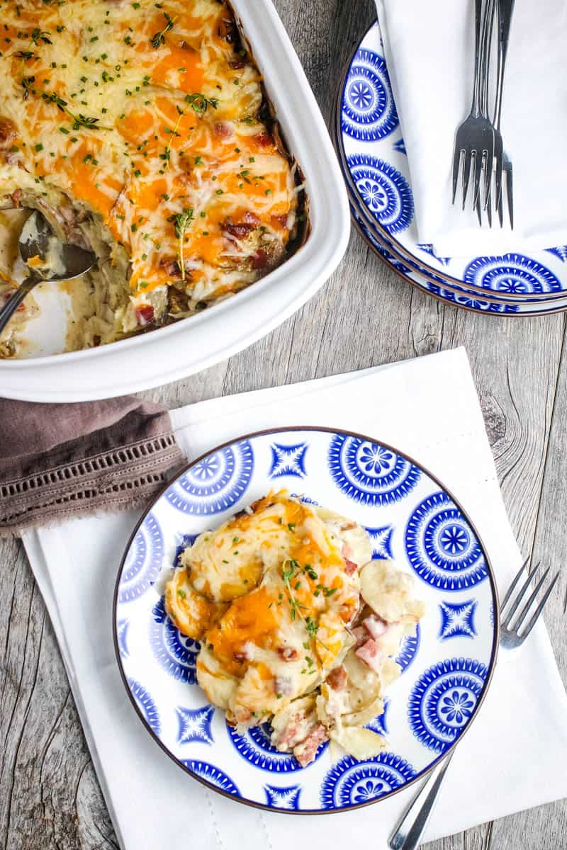 homemade scalloped potatoes with cheese on blue and white decorative plate on wood countertop with fork and entire casserole in white baking dish