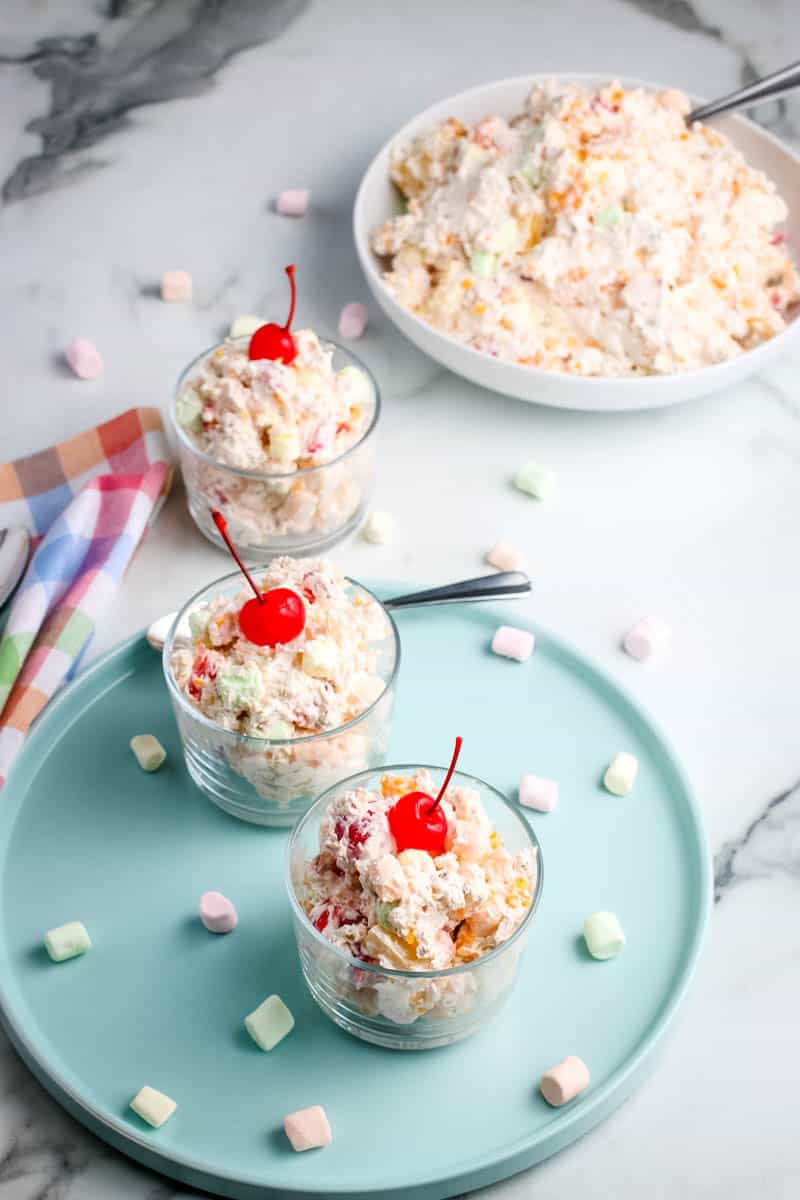 classic ambrosia salad recipe made in large bowl and placed in small glass serving bowls with cherries on top, on blue plate, on white marble countertop