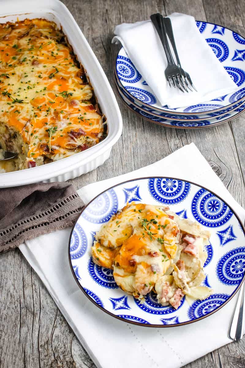 scoop of classic scalloped potatoes with cheese and chunks of ham on blue and white plate, on wood countertop with white and brown napkins