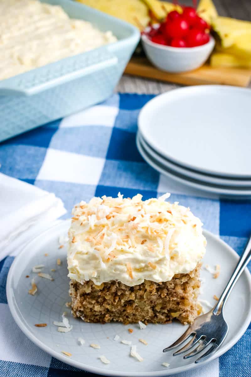 slice of easy pineapple cake, topped with toasted coconut, on gray plate with fork on blue checkered tablecloth