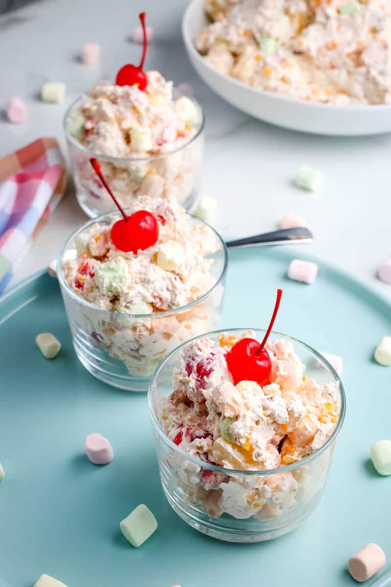 small bowls of marshmallow fruit salad topped with stemmed maraschino cherries, on blue plate with pastel colored mini marshmallows