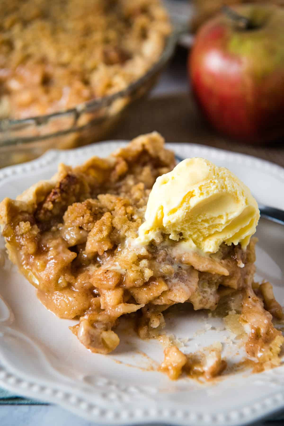 half eaten slice of apple pie with crumb topping, with scoop of vanilla ice cream on top, on white plate