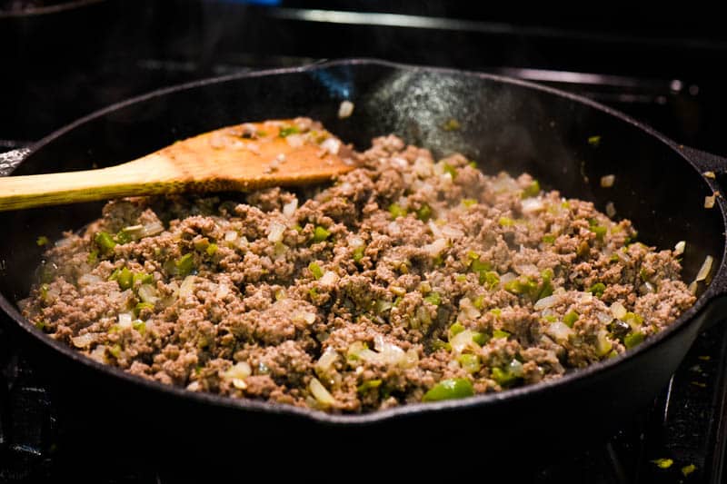 ground beef with peppers and onions and mushrooms, cooking in cast iron skillet with wooden spatula for stirring