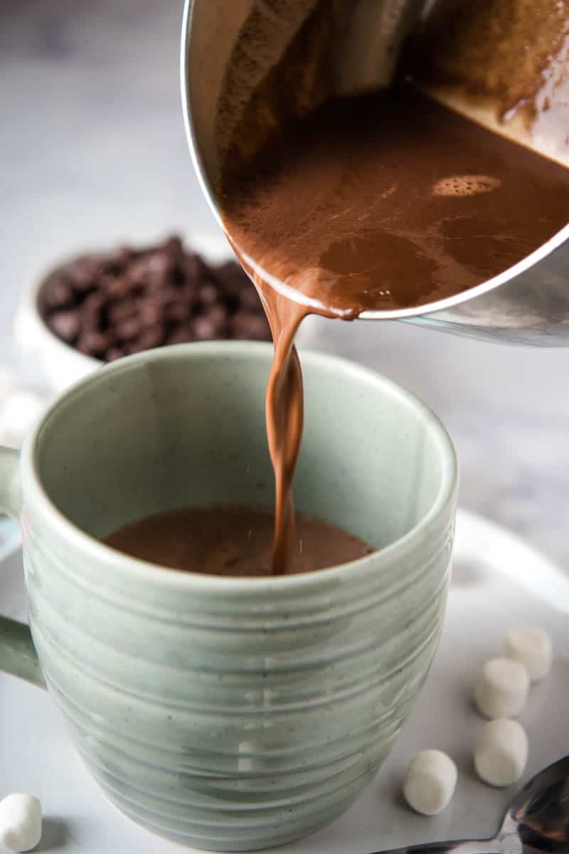 pouring creamy hot chocolate from saucepan into light blue-green cup, sitting on gray plate with mini marshmallows