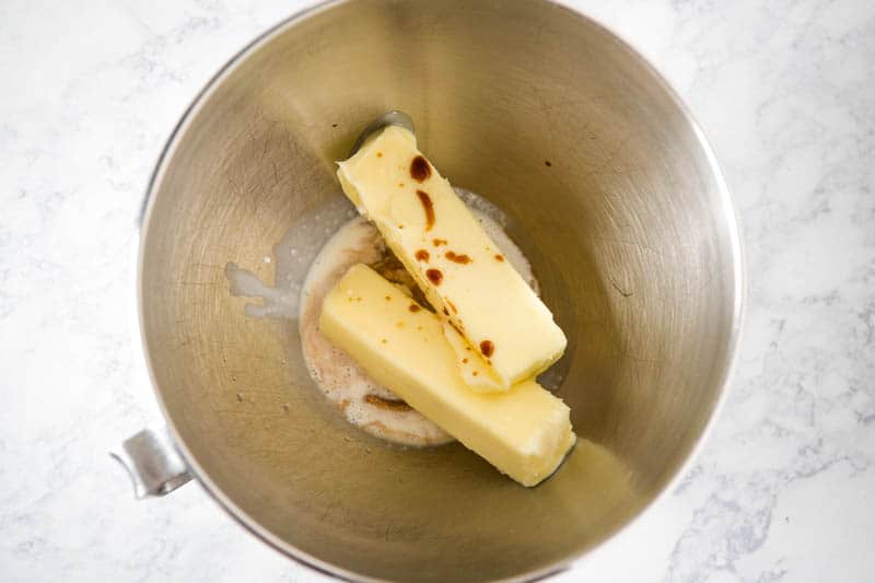 mixing together butter, vanilla extract, almond extract, and heavy whipping cream in KitchenAid mixing bowl on white marble countertop