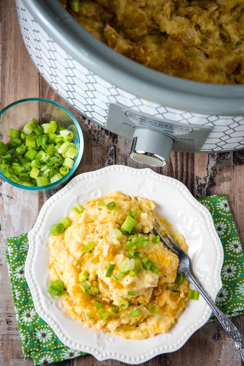 white plate full of CrockPot hash brown casserole with fork, with gray slow cooker and small bowl of green onions