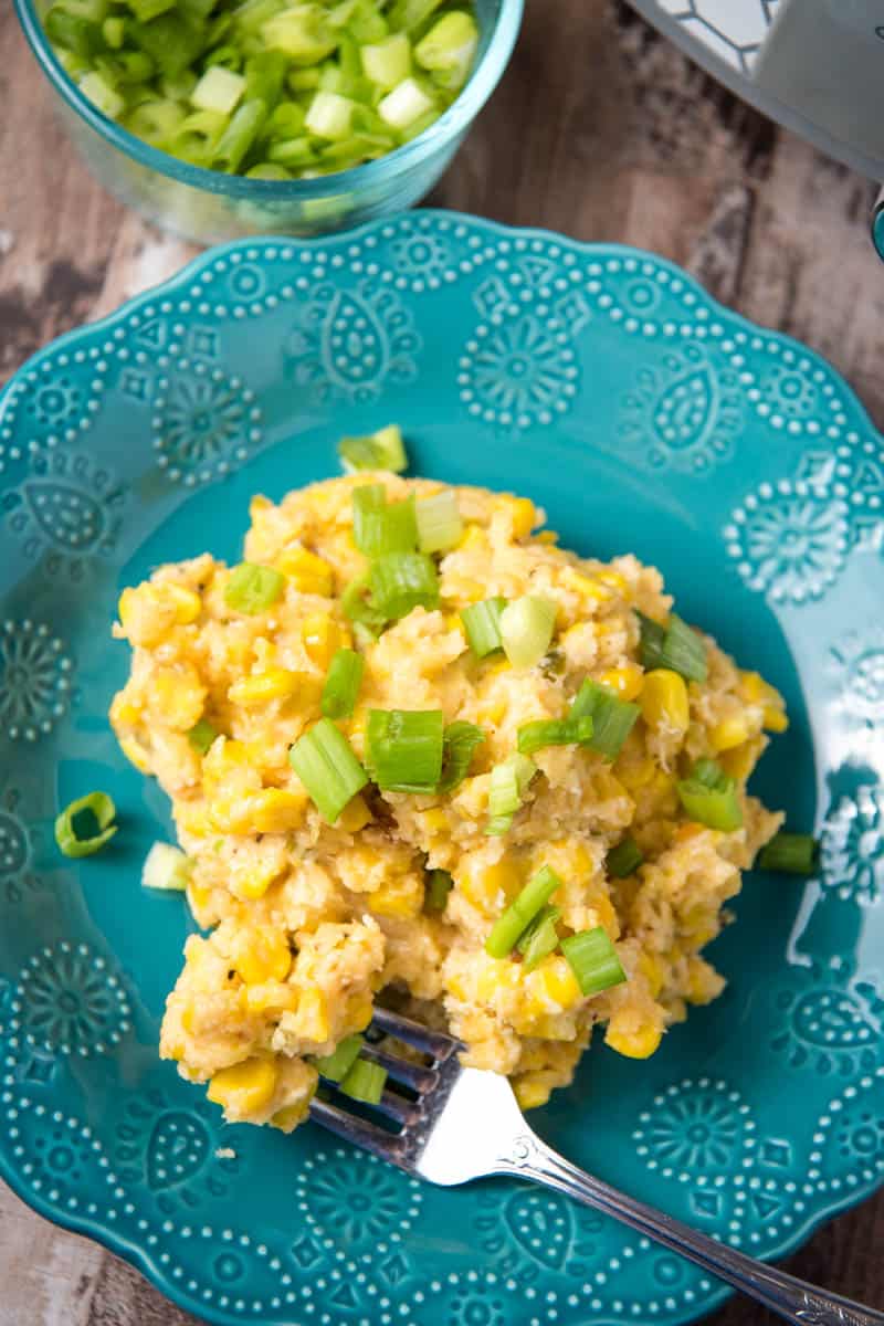 serving of slow cooker corn casserole, sprinkled with green onions, on teal plate with fork, on wooden countertop