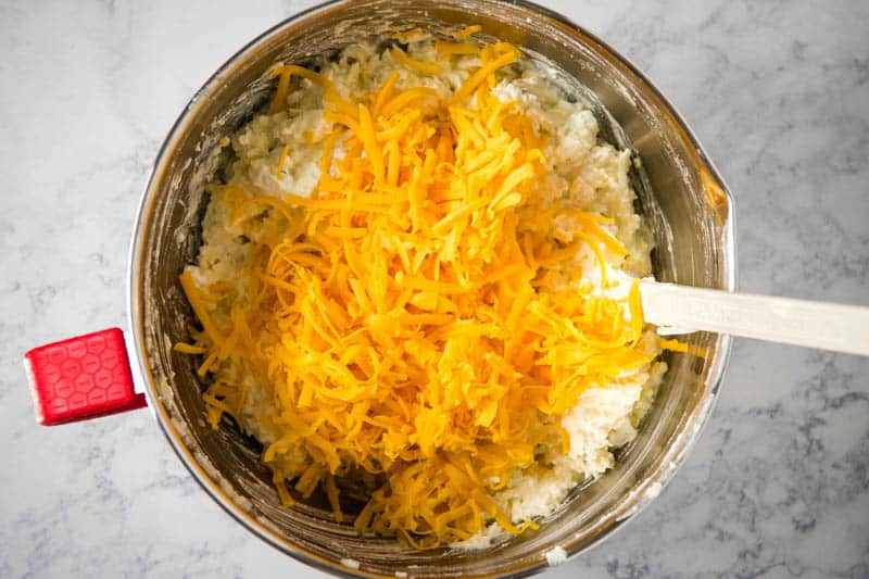 stirring cheddar cheese into hash brown mixture in metal mixing bowl with spatula