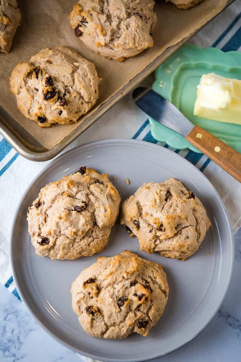 soda bread scones on gray plate with jadeite butter dish and butter knife, and baking sheet full of scones, on white marble countertop with blue and white striped kitchen towel