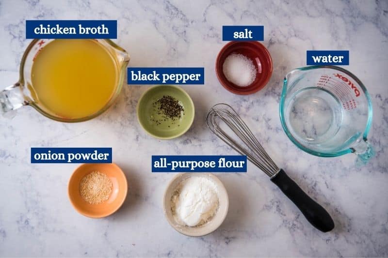 chicken gravy ingredients, including chicken broth from bouillon, black pepper, salt, water, onion powder, and all-purpose flour on white marble countertop with black whisk